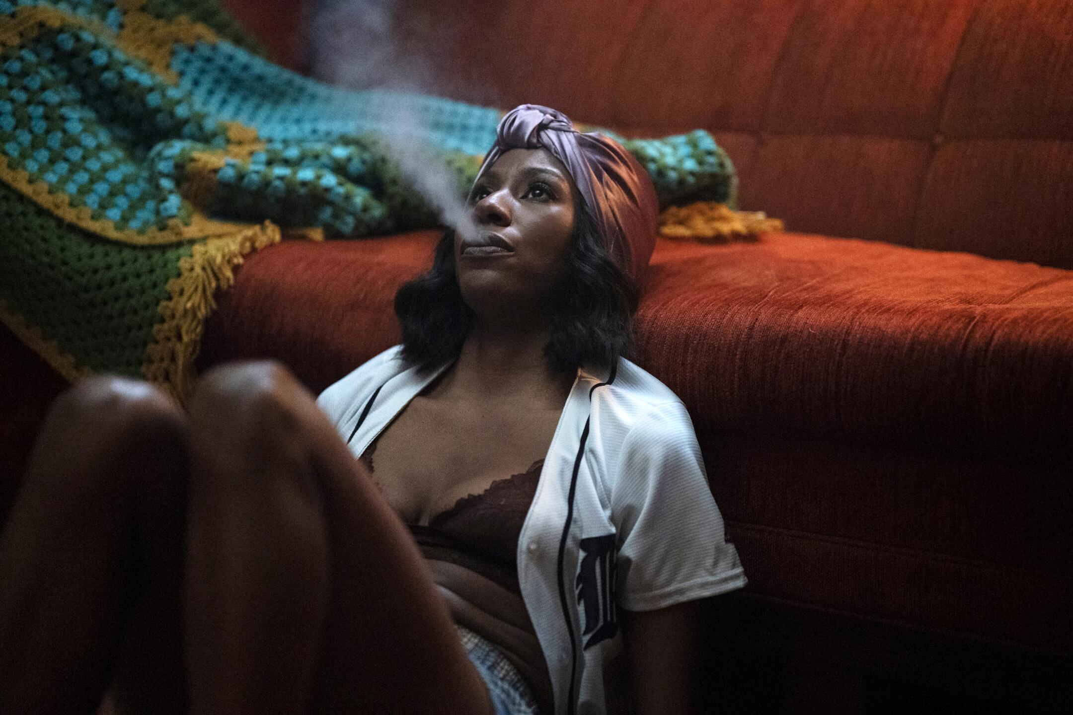 A woman sits on the floor, leaning against her couch exhaling smoke, in