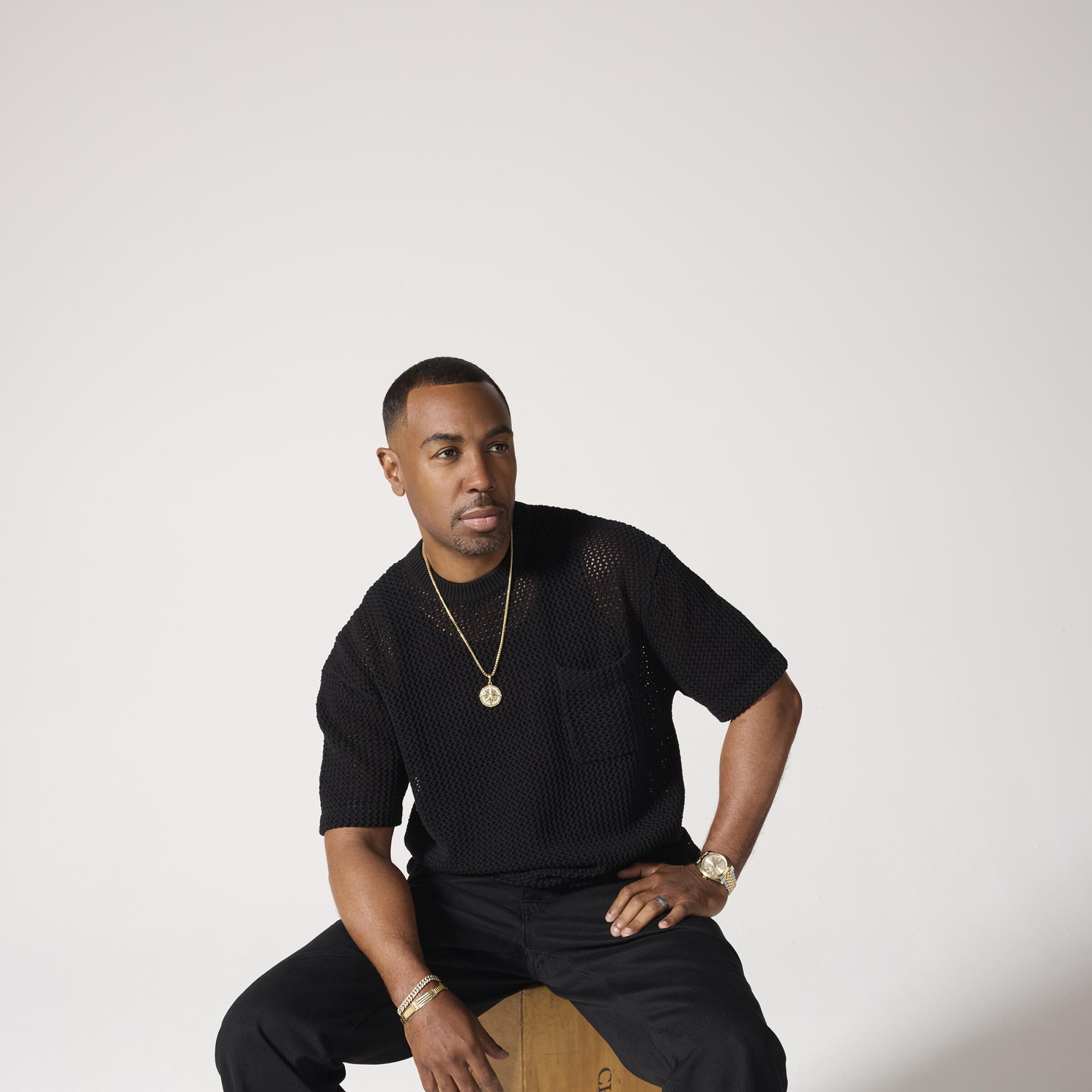 A man dressed in black sits on a block of wood for a portrait.