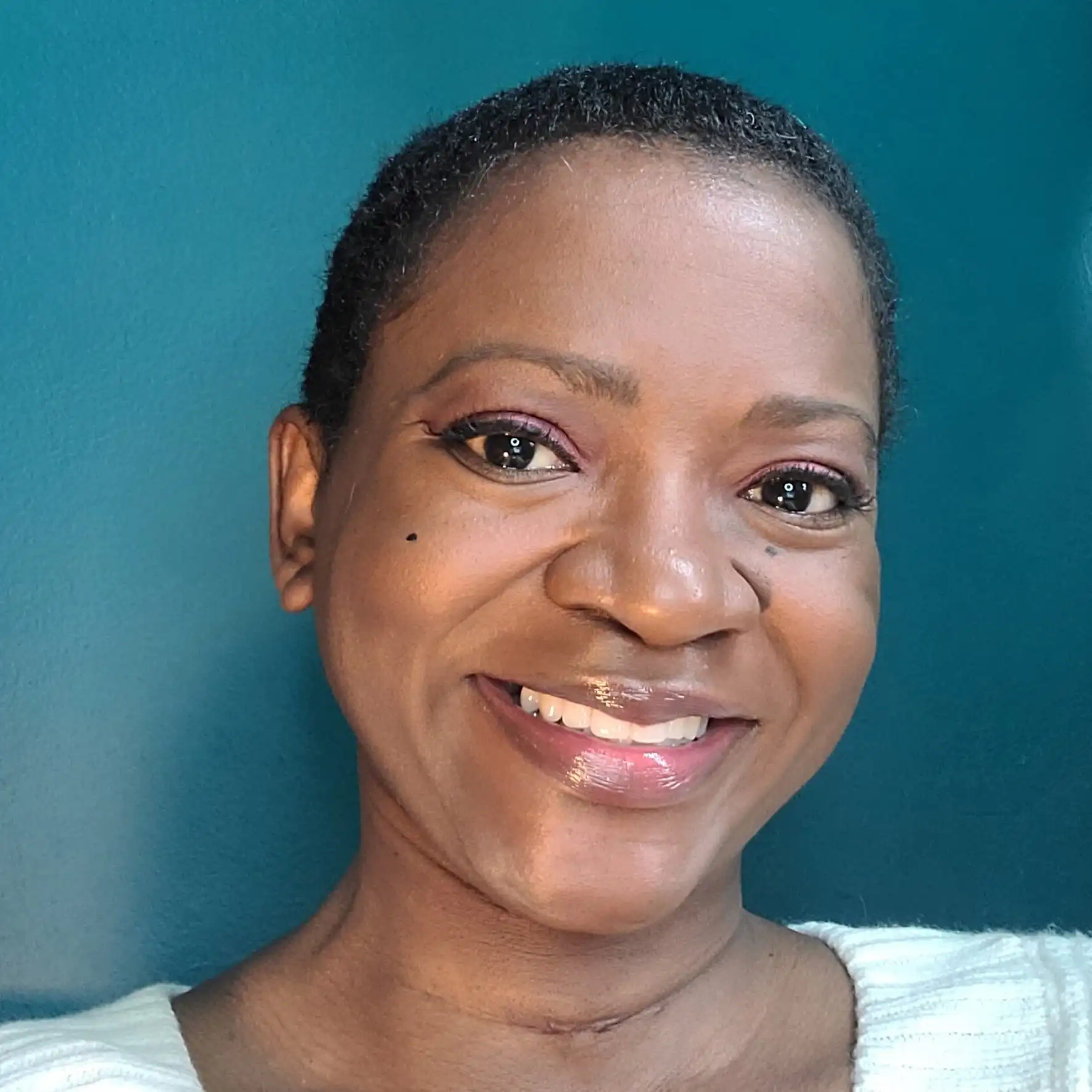 Photo of a Black female with dark brown complexion, close cropped black hair, wearing a wide smile, and white sweater, in front of a teal colored wall.