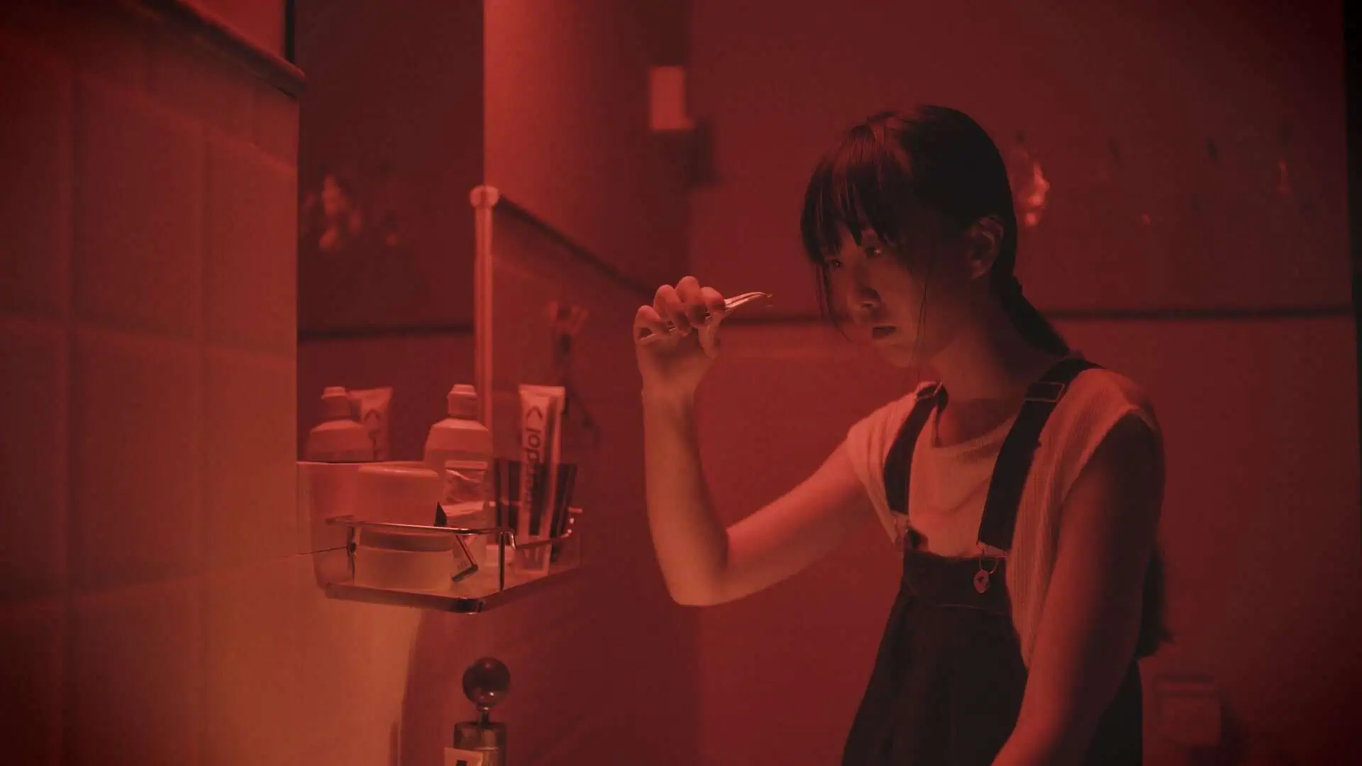 A young person in overalls holds up tweezers holding a contact lens, under red lighting, looking at it intently with a serious expression.