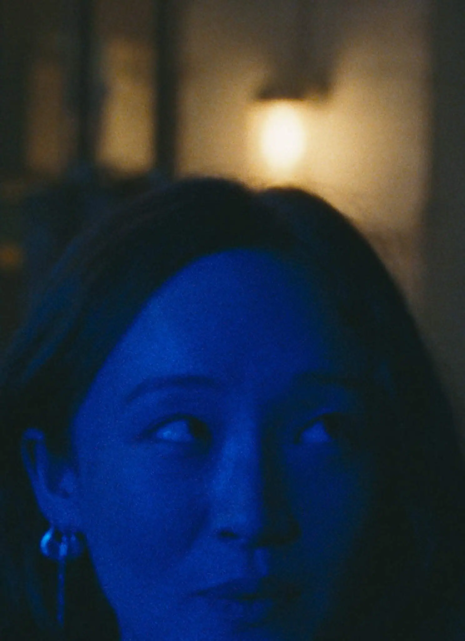 A woman and a man stand close together at night, illuminated by blue lighting. The woman looks at the man with a soft expression, while the man gazes intently at her. A blurred, warm light glows in the background.