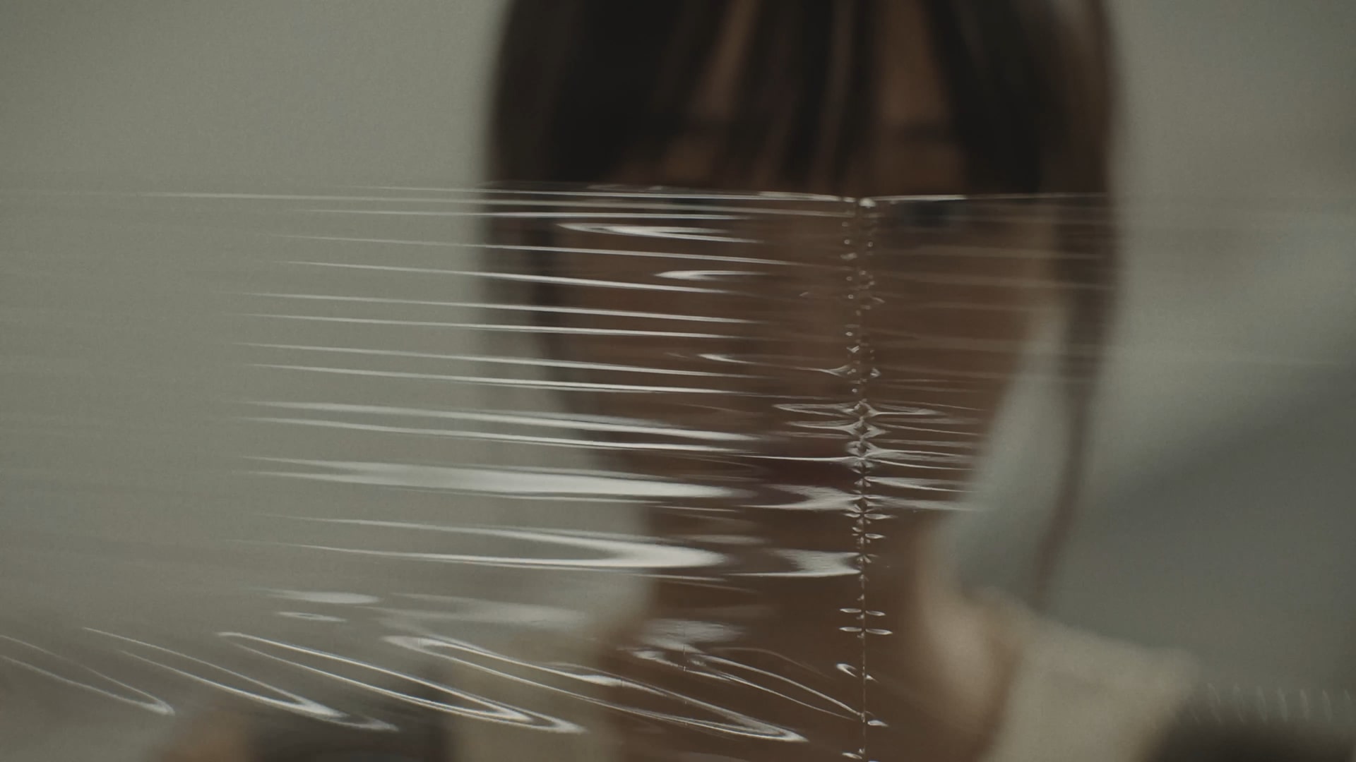 A person with blurred facial features is seen through a sheet of transparent, rippled plastic film, creating a distorted effect. The background is neutral and out of focus.