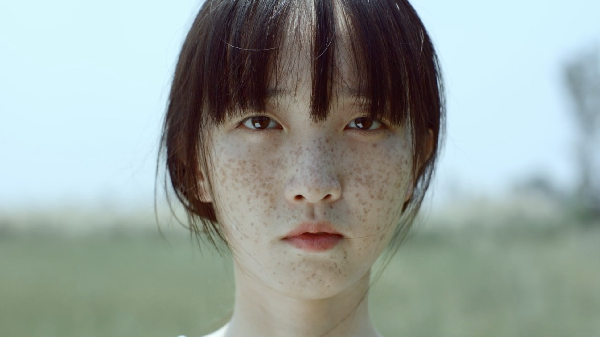 A young person with straight dark hair and freckles looks directly at the camera, standing outdoors with a blurred grassy background under bright daylight.