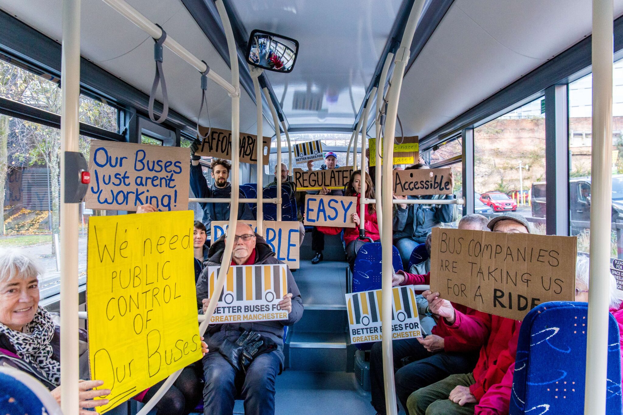 A group of passengers sat on a bus holding up placards stating 'We need public control of our buses', 'bus companies are taking us for a ride', 'better buses for Greater Manchester', 'EASY', 'Acessible', 'AFFORDABLE