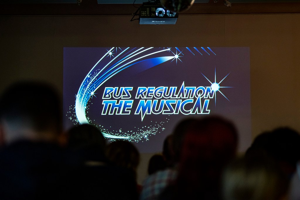 A projected logo that states 'Bus Regulation: the Musical' in front of a seated audience