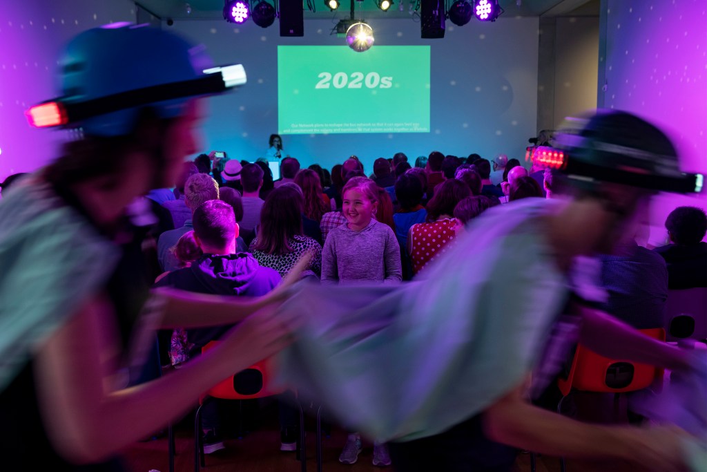 Two performers on rollerskates skate past a seated crowd with a child stood watching them smiling. In the background is a projected screen stating '2020s'.