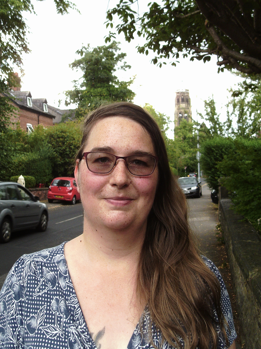 A head and shoulders portrait of Cazz Blase stood at the side of the road with trees, parked cars and a church tower in the background.