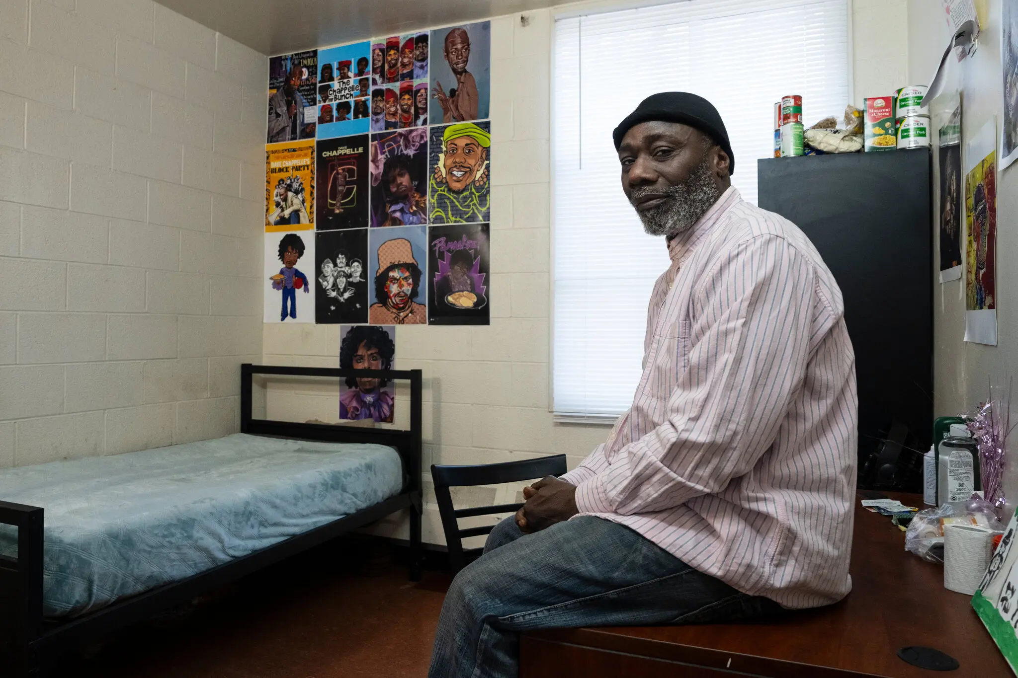 A man sits on a desk in a room that has a single bed and a window.