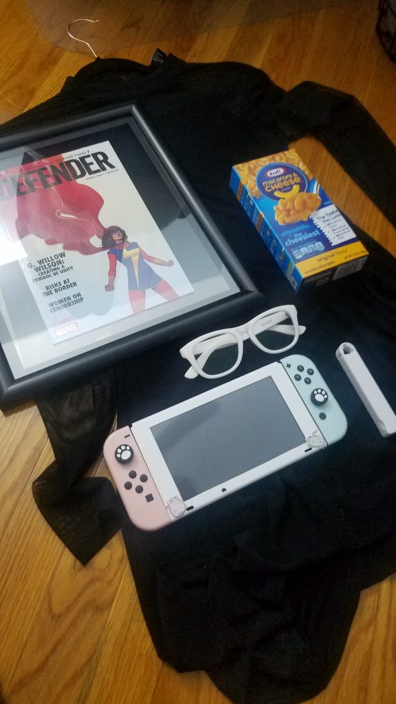 A photo of a pile of miscellaneous items sitting on a wooden floor: A framed comic issue: a box of Kraft Macaroni & Cheese, a Nintendo Switch, a pair of thick, white framed glasses, and a small, white container.