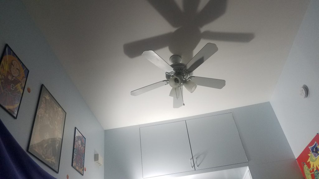 A photo of a low angle view of a ceiling fan in a dimly-lit room. Several posters hang around the surrounding walls.