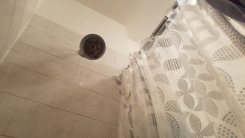 A photo of a low angle view of a shower head. A shower curtains hangs on a rod adjacent to the shower head.