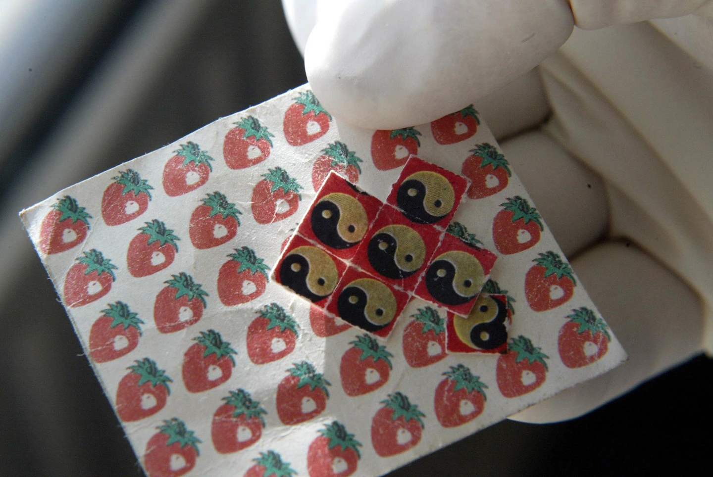 A gloved hand holds two sheets of LSD tabs, one designed to look like strawberries and one designed to look like yin and yang symbols