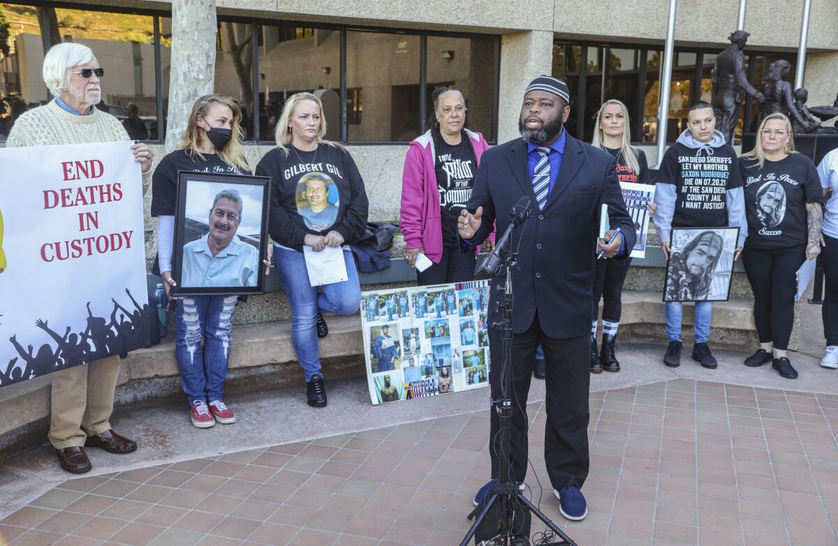 A man wearing a suit and a taqiyah speaks into a microphone, flanked by people holding portraits of loved ones and posters.
