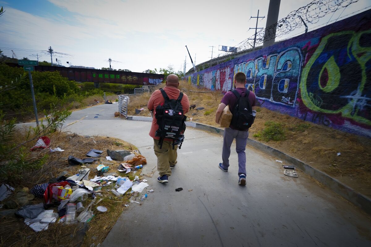 Two men in backpacks walk down a trash-strewn road alongside a barbed wire-topped wall toward a riverbed.