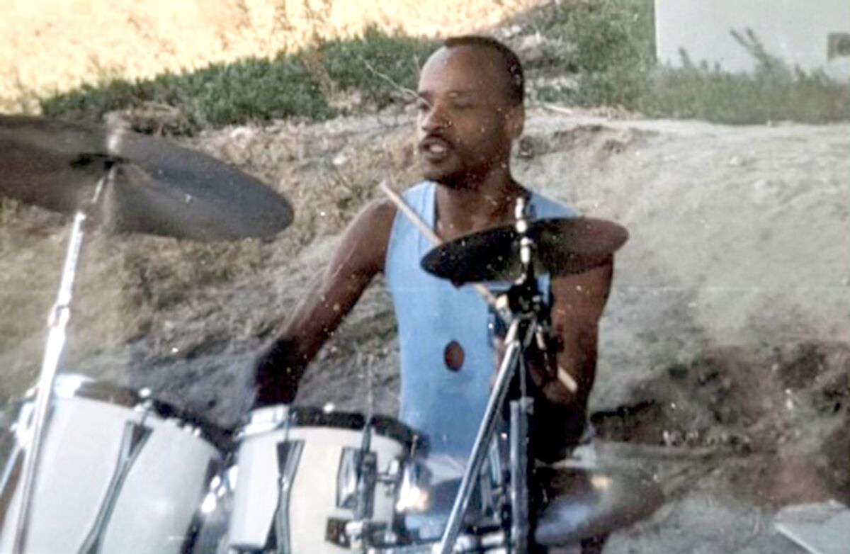 A thin man wearing a sleeveless shirt plays a drum kit in a backyard.