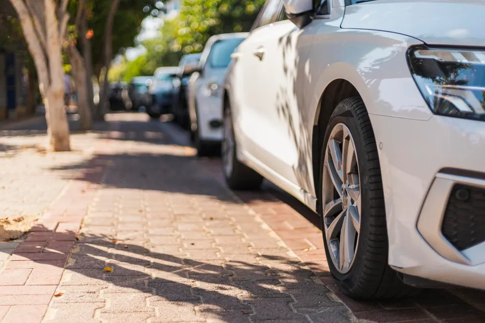A white vehicle is parked close to the sidewalk on a warm sunny day. Trees provide shade along the street, where additional cars are visible in the background
