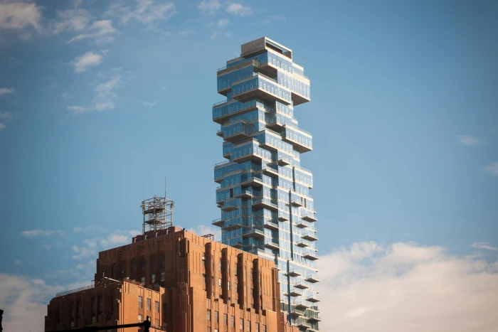 A tall, uniquely designed skyscraper with a stacked, offset glass balcony structure stands next to a red-brick building with art deco elements