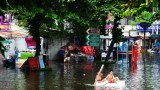 A flooded Bangkok street with shops and boys paddling on a container.