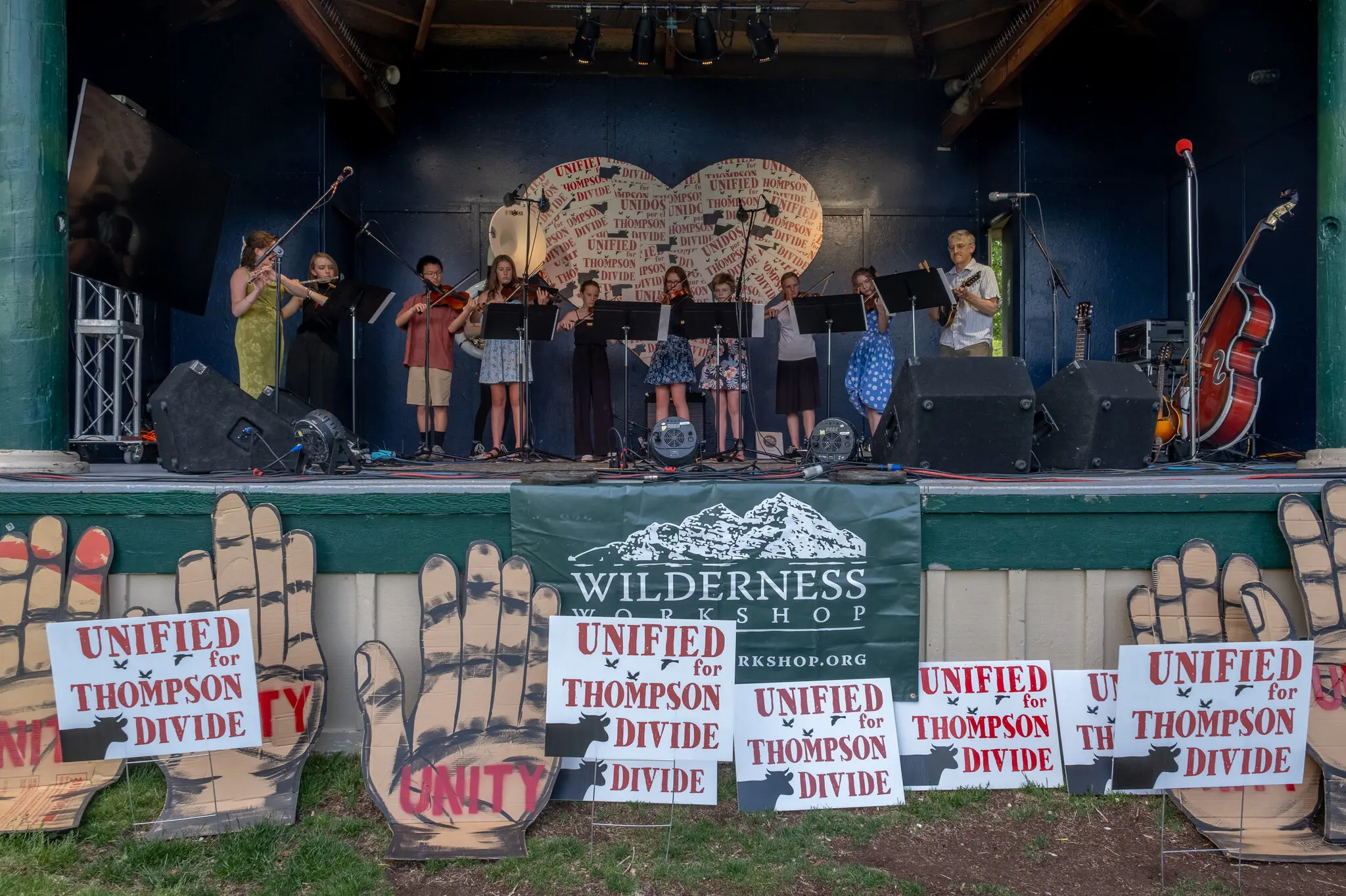 Children lined up on a green outdoor stage with two adults. One child plays a flute. The others have violins or violas. Placards at the base of the stage read “Unified for Thompson Divide.”