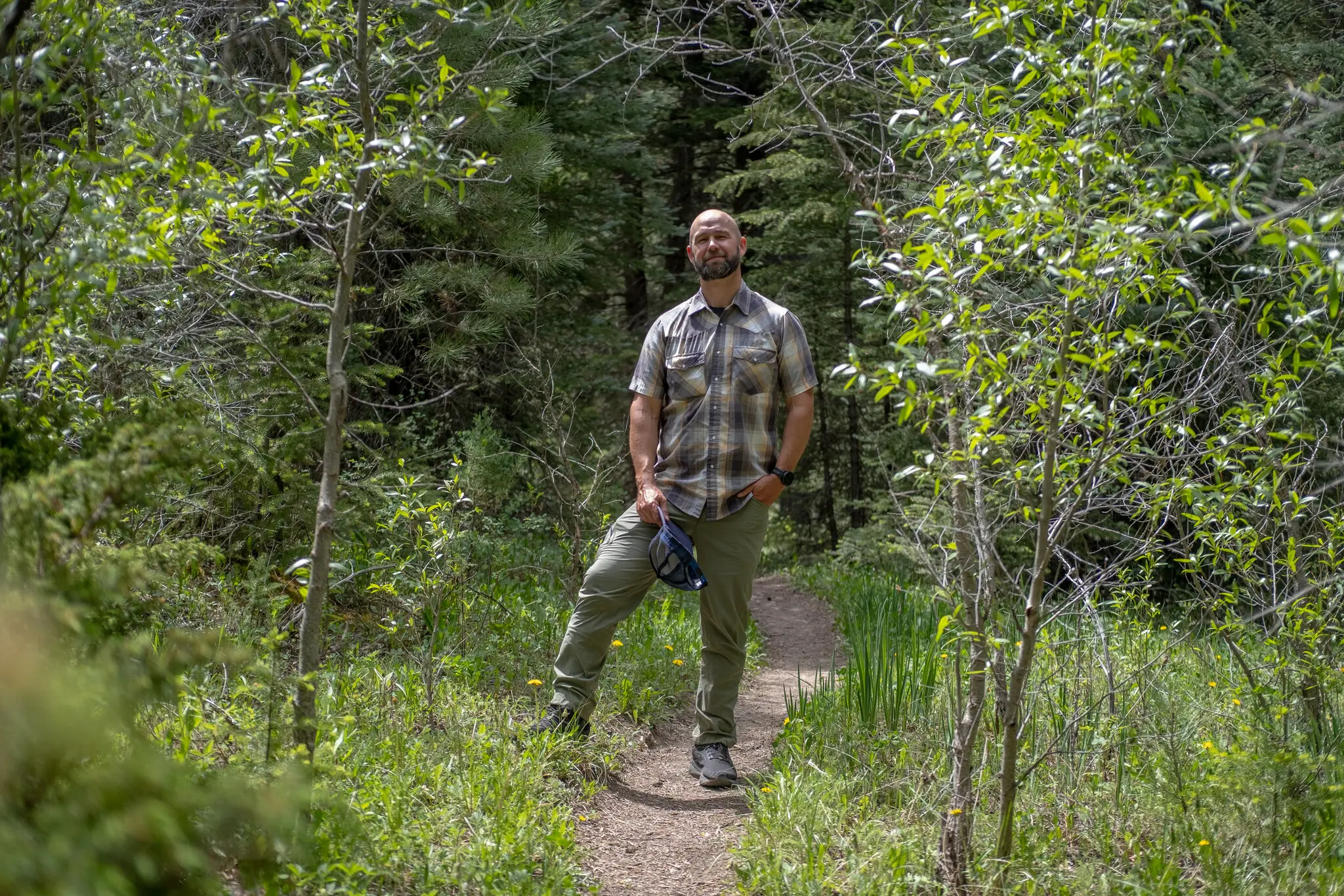 Peter Hart stands at a forest trail. He has a beard and a brown plaid shirt and holds a baseball cap in his right hand.