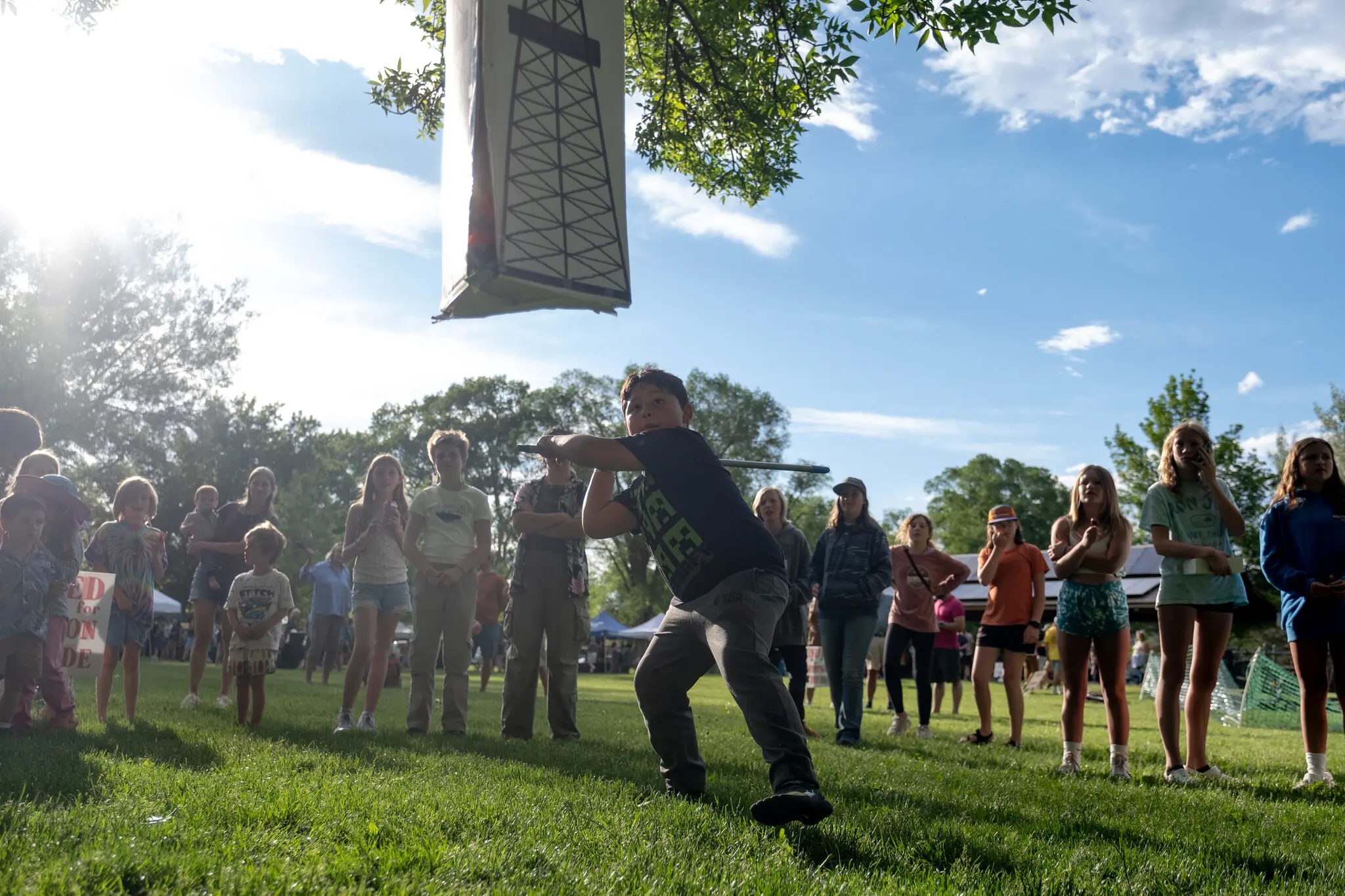 A child aiming a long stick at a piñata hanging from a tree branch. The piñata has an oil rig pictured on the side. Dozens of other children and a few adults are watching.