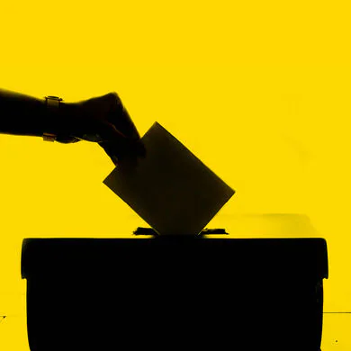 Black silhouette image in front of yellow background of a hand dropping a vote into a ballot