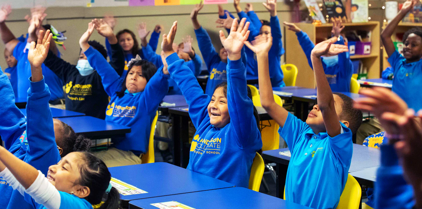 Elementary school students sit at their desks with blue sweatshirts, smiling and raising their hands